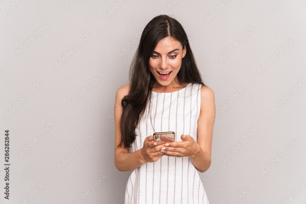 Image of pretty brunette woman wearing dress smiling while holding modern smartphone Image of pretty brunette woman wearing dress smiling while holding modern smartphone