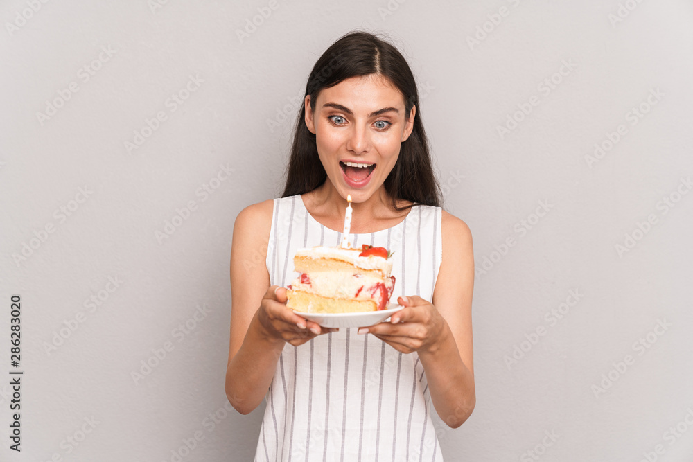 Image of happy brunette woman wearing dress smiling and holding birthday cake with candle Image of happy brunette woman wearing dress smiling and holding birthday cake with candle