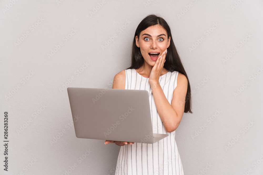 Image of excited brunette woman wearing dress smiling while holding silver laptop computer Image of excited brunette woman wearing dress smiling while holding silver laptop computer