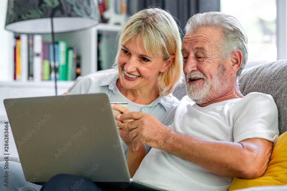 Senior couple using laptop in living room. White man and woman sitting on couch looking at laptop. Interesting talk. Senior couple using laptop in living room. White man and woman sitting on couch looking at laptop. Interesting talk.