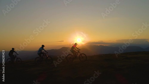 AERIAL, SILHOUETTE, LENS FLARE: Breathtaking evening sunbeams shine on the tourists riding ebikes in the scenic nature. Three downhill bikers pedalling their bicycles up along grassy hill at sunrise.