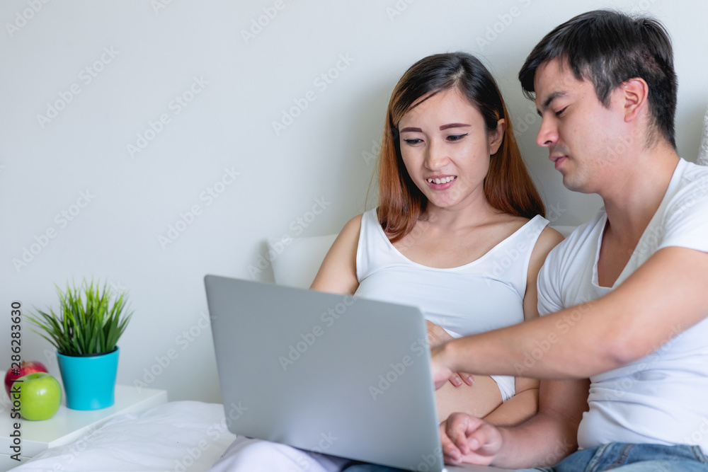 Pregnant woman in bed with man. Asian woman with white man. Using laptop. Mix race relationship concept. Happy smile with copy space.