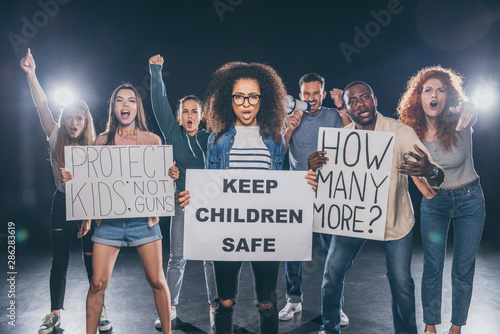 emotional african american woman holding placard with keep children safe lettering near group of people on black © LIGHTFIELD STUDIOS