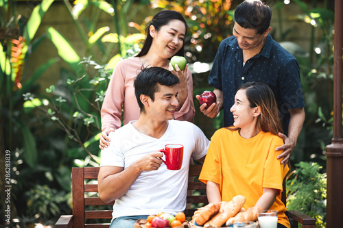 Family lunch outdoor. Married mix race couple with grand parents. White man, Asian pregnant woman with senior asian couple in tropical garden. Talking to eath other, laughing.