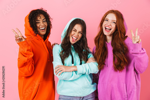 Pleased smiling young three multiethnic girls friends posing isolated over pink wall background showing peace gesture.