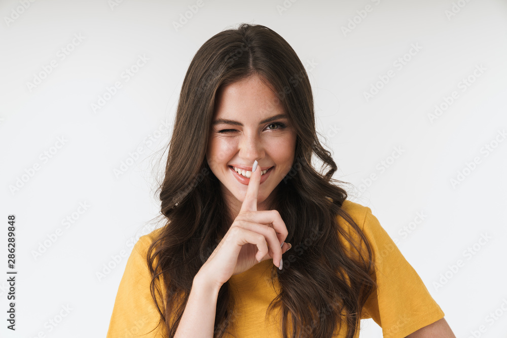 Image of charming brunette woman wearing casual t-shirt holding index finger on lips Image of charming brunette woman wearing casual t-shirt holding index finger on lips