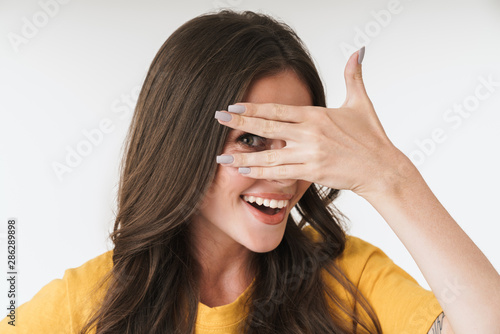 Image of alluring brunette woman wearing casual t-shirt smiling and peeking behind her hand
