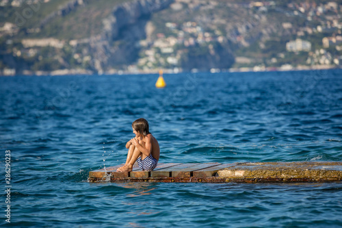 Little boy, child, sitting on dock in the sea, enjoying the view while waves splashing around © Tomsickova
