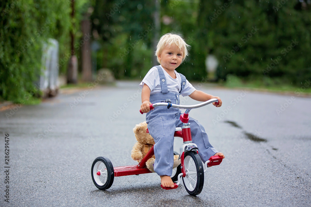 Cute little boy, with teddy bear toy, riding tricycle on the street in the rain, barefeet Cute little boy, with teddy bear toy, riding tricycle on the street in the rain, barefeet
