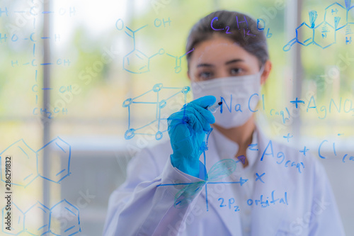 A young beautiful asian girl scientist wear health mask writing table of the elements on glass board in laboratory at morning classroom. Preparing for new biologic lab. Selective focus on woman face.