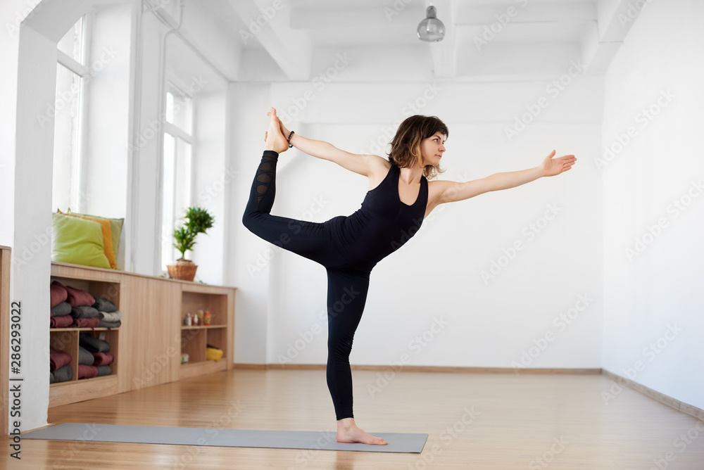 Side portrait view of beautiful young woman wearing black tank top and leggings, workout alone against white wall, doing yoga or pilates exercise. Standing in Lord of dance pose. Full length