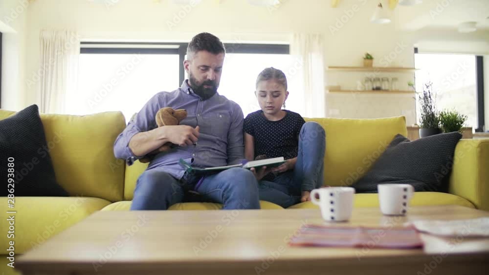 Mature father with small daughter sitting on sofa indoors, reading a book. Mature father with small daughter sitting on sofa indoors, reading a book.