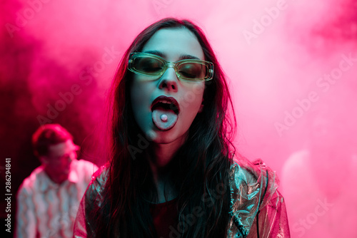 beautiful girl in sunglasses with lsd on tongue in nightclub with pink smoke photo