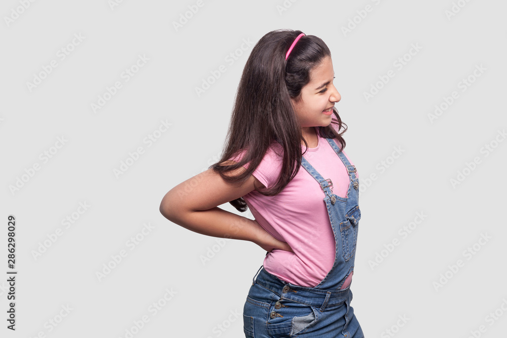 Spine back pain. Profile side view Portrait of sick brunette young girl in pink t-shirt and blue overalls standing and holding her painful back. indoor studio shot, isolated on light gray background. Spine back pain. Profile side view Portrait of sick brunette young girl in pink t-shirt and blue overalls standing and holding her painful back. indoor studio shot, isolated on light gray background.