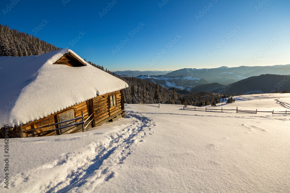 Human footprint path in white deep snow leading to small old wooden forsaken shepherd hut in mountain valley, spruce forest, woody dark hills, bright sun on clear blue sky copy space background. Human footprint path in white deep snow leading to small old wooden forsaken shepherd hut in mountain valley, spruce forest, woody dark hills, bright sun on clear blue sky copy space background.