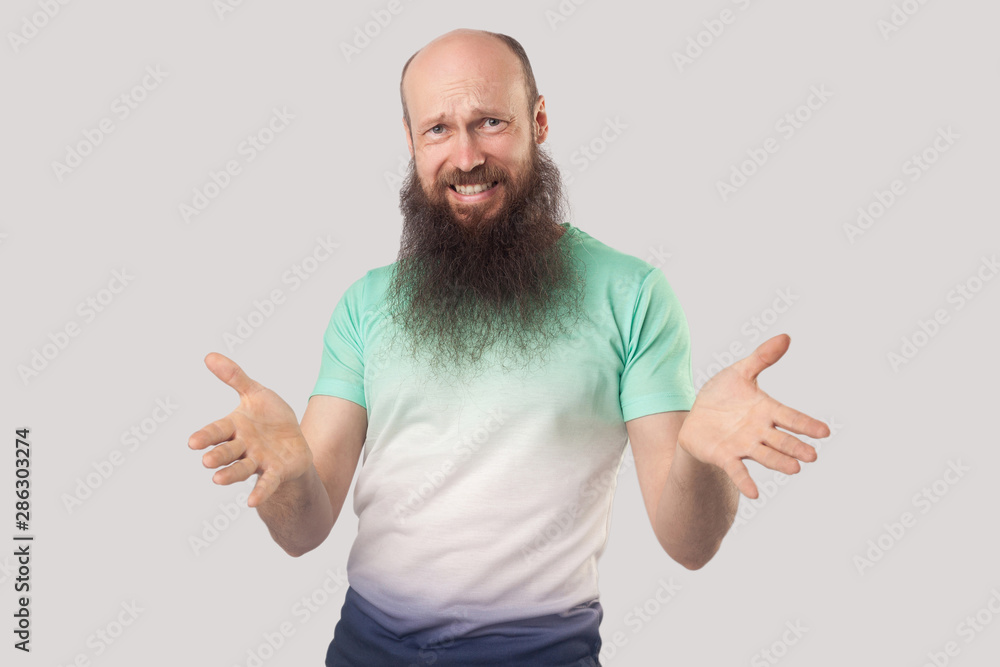 What do you need? Portrait of angry middle aged bald man with long beard in light green t-shirt standing, looking at camera with raised arms and asking. indoor studio shot isolated on grey background What do you need? Portrait of angry middle aged bald man with long beard in light green t-shirt standing, looking at camera with raised arms and asking. indoor studio shot isolated on grey background