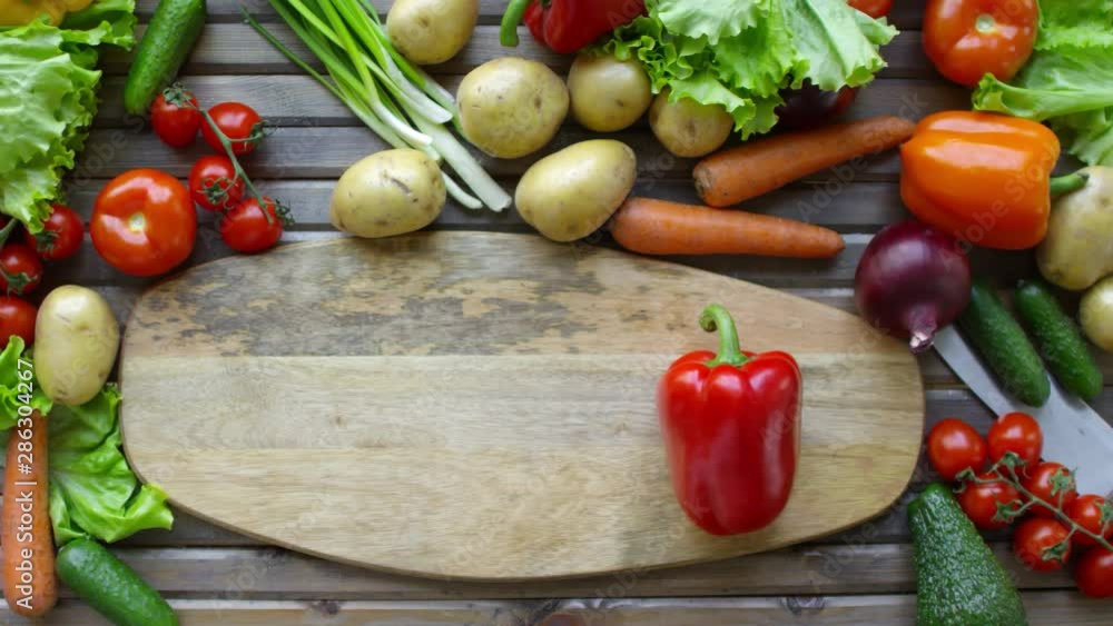 Top view of hands of unrecognizable male cook spinning red bell pepper on wooden cutting board with copy space on kitchen table with lots of fresh vegetables around Top view of hands of unrecognizable male cook spinning red bell pepper on wooden cutting board with copy space on kitchen table with lots of fresh vegetables around