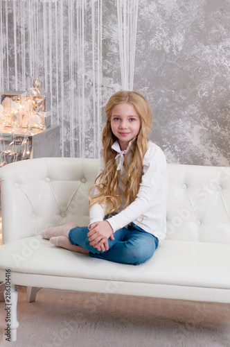 Christmas. Beautiful blonde girl child teenager smiling sitting on a white sofa in a room decorated with Christmas garland