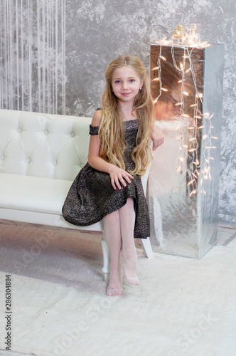 Blonde, beautiful girl in a black dress sitting on a white sofa near the Christmas garland