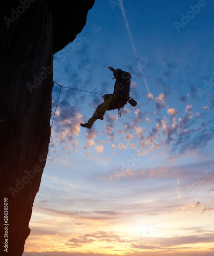 Climber silhouette rappelling from summit of challenging cliff route. Risky extreme jumping flight in air. Magical blue sunrise sky with copy space. Low angle view. Freedom and courage concept
