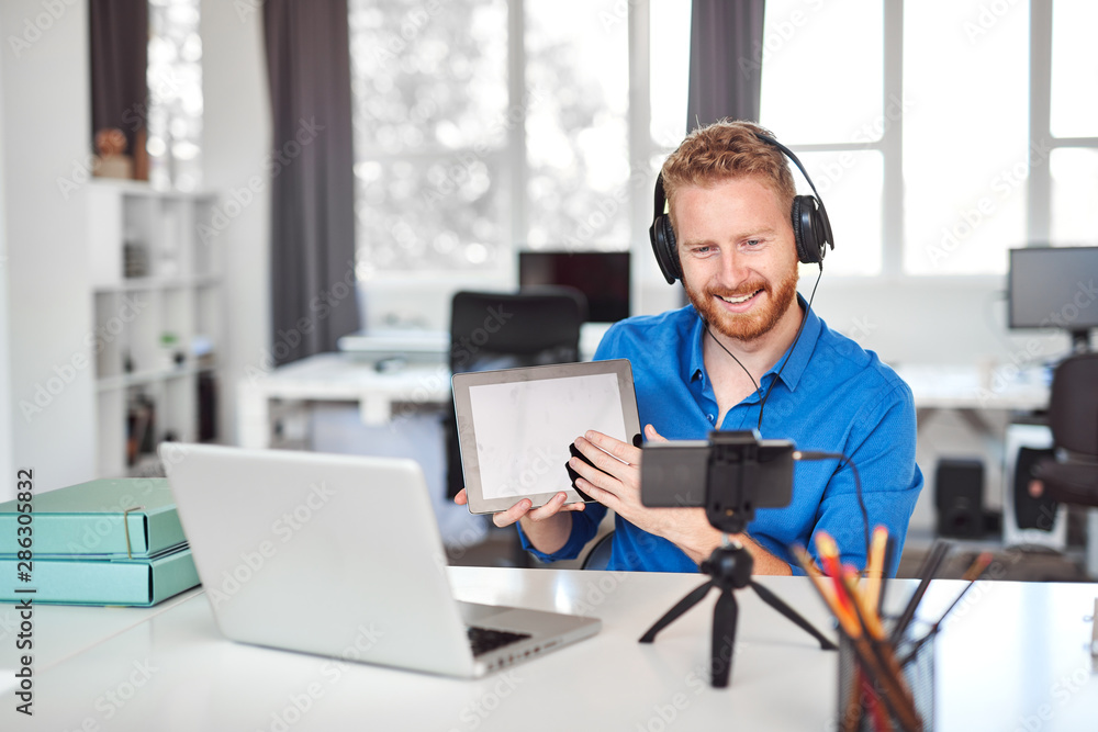 Young Caucasian hardworking employee havining video call over smart phone. In hands is tablet and on head are headphones. Remote business concept. Young Caucasian hardworking employee havining video call over smart phone. In hands is tablet and on head are headphones. Remote business concept.
