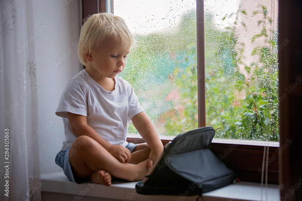 Adorable little boy, sitting on window shield, playing on tablet on a rainy day Adorable little boy, sitting on window shield, playing on tablet on a rainy day