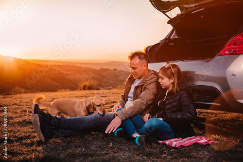 Father and daughter with dog sitting by the car on the hill