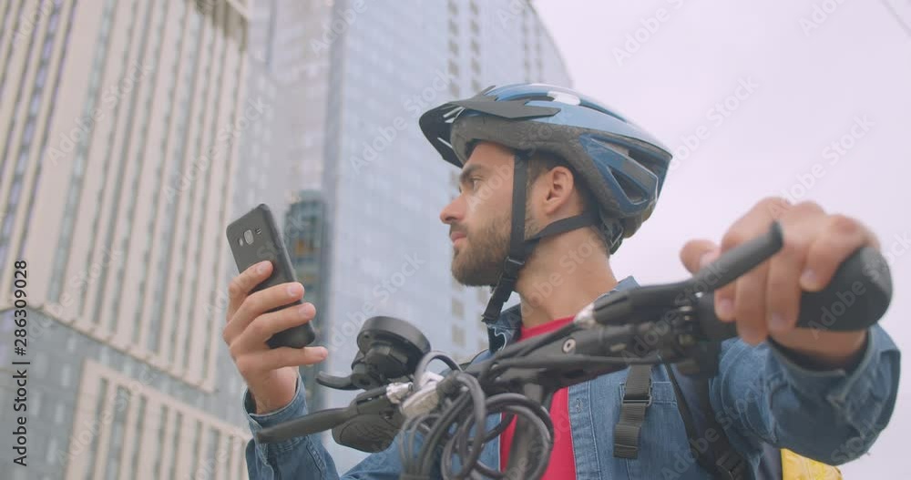 Closeup portrait of adult attractive caucasian delivery man standing with kick scooter and back pack using navigation app on phone in city Closeup portrait of adult attractive caucasian delivery man standing with kick scooter and back pack using navigation app on phone in city