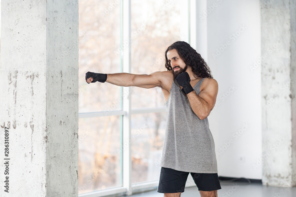 Portrait of angry muscular young adult man with curly long hair standing  and preparing to fight wall with fist in black gloves, showing professional  technique. Indoor, window background, martial arts Stock Photo |