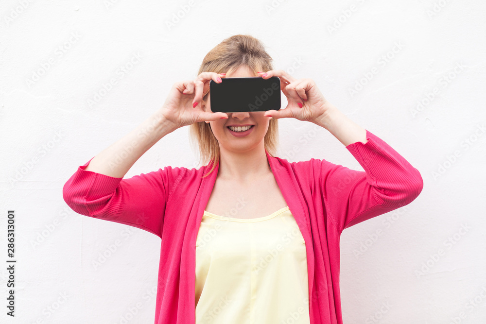 Cant see you! Portrait of happy modern young woman in pink blouse standing, holding and covering eyes with smartphone, like eyeglasses and toothy smile. Indoor, isolated, studio shot, white background Cant see you! Portrait of happy modern young woman in pink blouse standing, holding and covering eyes with smartphone, like eyeglasses and toothy smile. Indoor, isolated, studio shot, white background