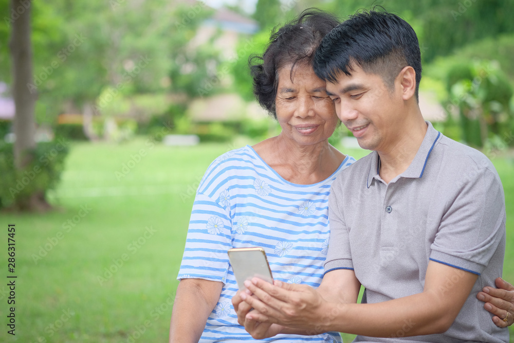 Middle-aged Asian mother and son Looking at a smartphone with a smile and being happy at the park Is an impressive warmth Middle-aged Asian mother and son Looking at a smartphone with a smile and being happy at the park Is an impressive warmth
