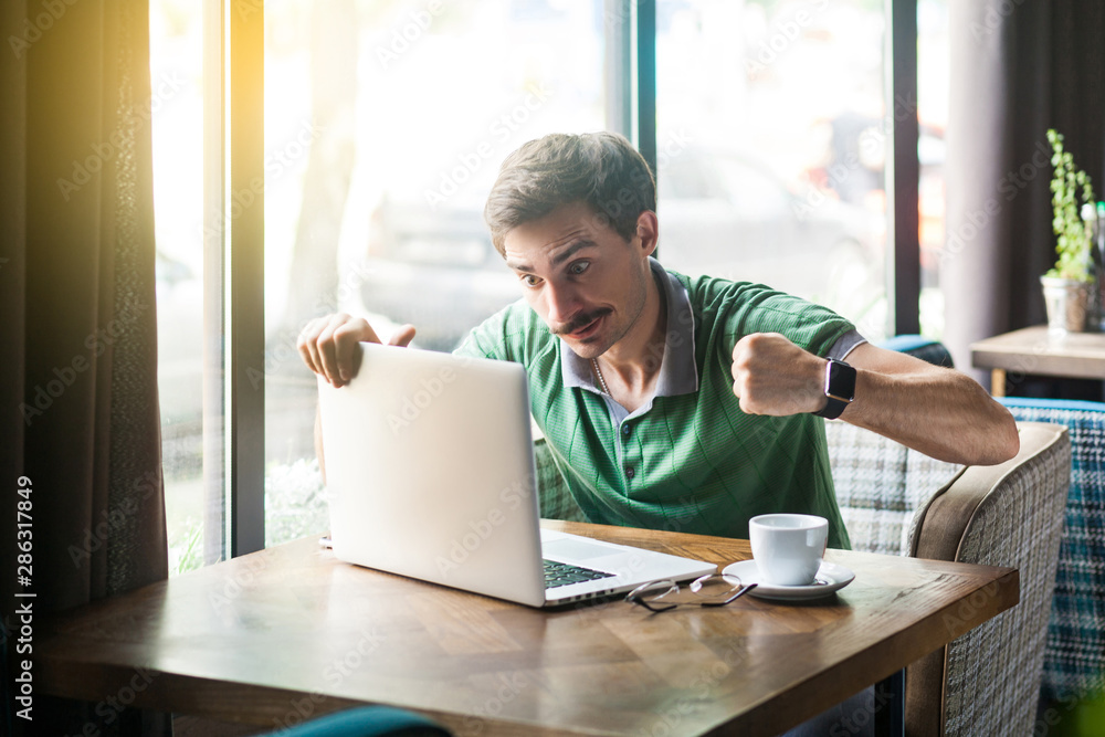 Young angry businessman in green t-shirt sitting with aggressive face and boxing fists and try to punching his laptop. problem on business and freelancing concept. indoor, near big window at daytime. Young angry businessman in green t-shirt sitting with aggressive face and boxing fists and try to punching his laptop. problem on business and freelancing concept. indoor, near big window at daytime.