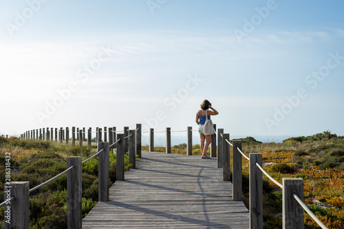 Woman from the back in a distance standing on a wooden walkway, contemplating the blue sky