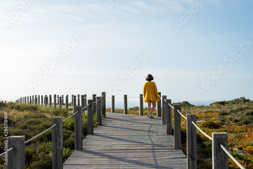 Woman from the back with a yellow jacket in a distance standing on a wooden walkway, contemplating the blue sky
