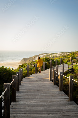 Fototapeta Naklejka Na Ścianę i Meble -  Woman in a distance standing in a wooden pathway looking at the beach below. Surrounded by vegetation and the sea.