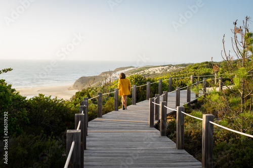Woman in a distance standing in a wooden pathway looking at the beach below. Surrounded by vegetation and the sea.