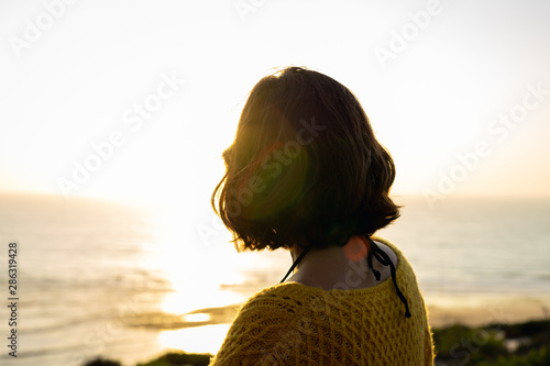 Girl with short hair and a yellow sweater watching the sunset on the beach below.