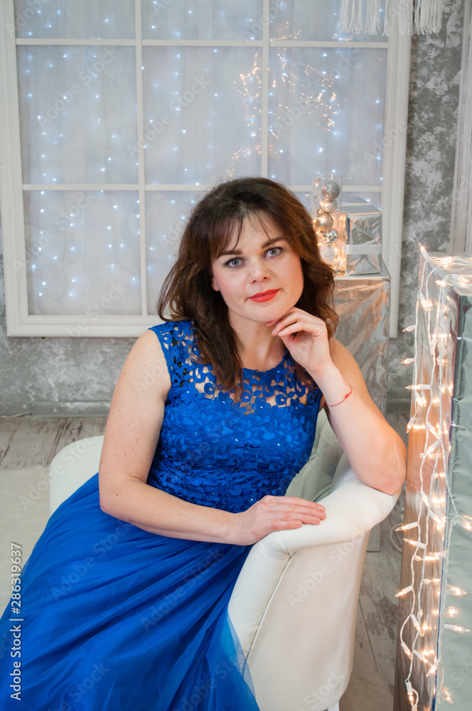 Fototapeta premium A very beautiful brown-haired woman with curly hair in a blue evening dress is sitting on a business sofa near the New Year's garland. Portrait, vertical format