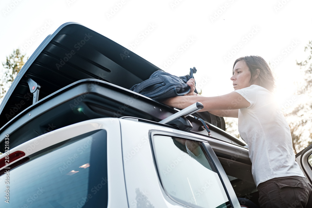A woman puts things in the roof rack of a car or in a cargo box, before a family trip on vacation, against the sky and trees, on a summer evening.