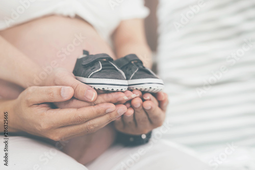 pregnant woman and her husband hand splice the tummy holding small shoes for the unborn baby in the belly relaxing at home in bedroom. wait birth date expecting a baby stand in the outdoors.