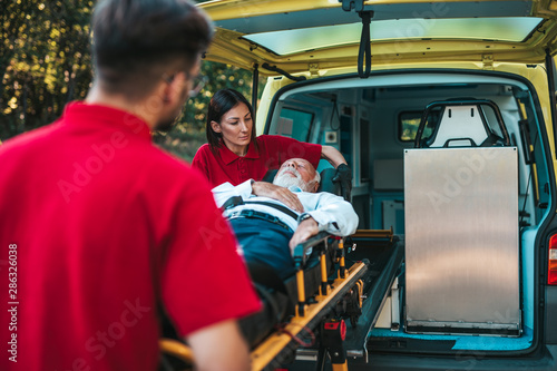 Emergency medical service at work. Paramedic is pulling stretcher with senior man with serious heart attack to the ambulance car. Help on the road. Drivers assistance concept.