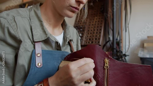 Pan shot of female tanner with blonde hair holding leather piece in wooden vice and stitching it with brown thread and two big needles