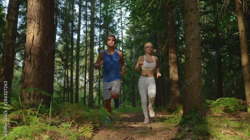 SLOW MOTION, LOW ANGLE, CLOSE UP: Fit couple exploring the peaceful forest during a relaxing jog on a sunny summer afternoon. Happy woman and man exercising by going trail running in the pine woods. SLOW MOTION, LOW ANGLE, CLOSE UP: Fit couple exploring the peaceful forest during a relaxing jog on a sunny summer afternoon. Happy woman and man exercising by going trail running in the pine woods.