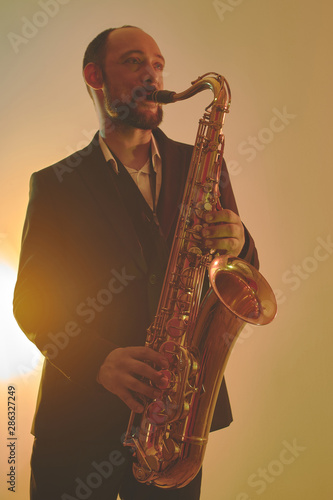 Portrait of professional musician saxophonist man in suit plays jazz music on saxophone, yellow background in a photo studio