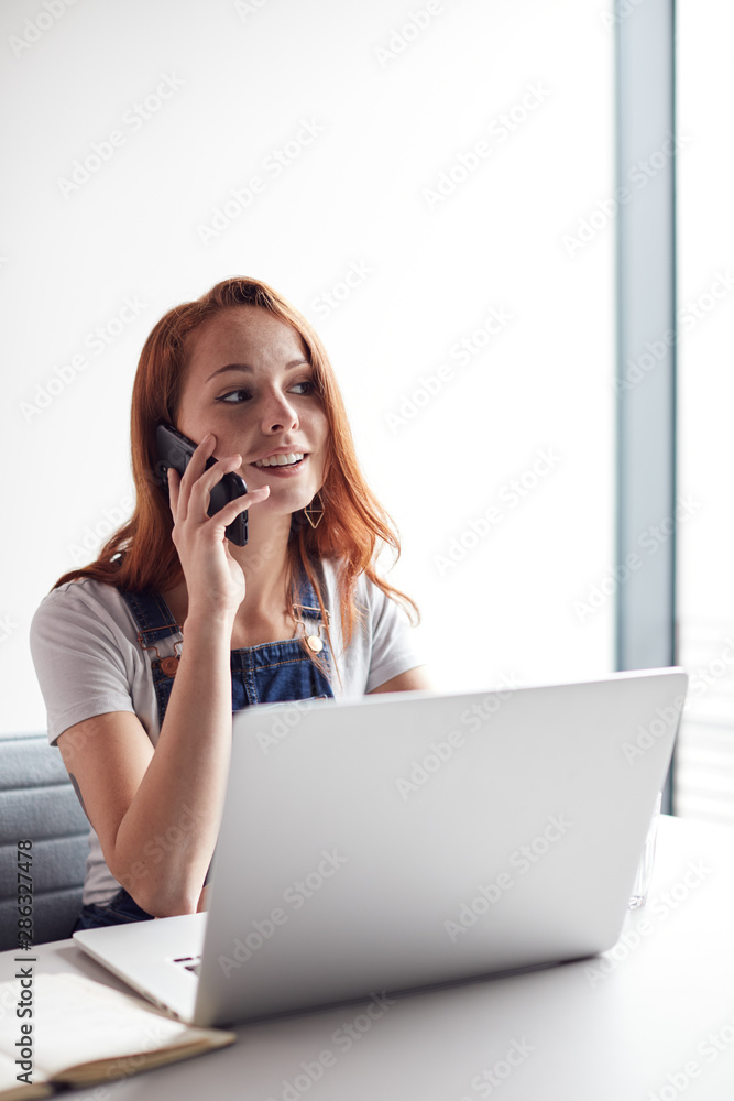 Casually Dressed Young Businesswoman Working On Laptop At Desk In Modern Workplace Making Phone Call Casually Dressed Young Businesswoman Working On Laptop At Desk In Modern Workplace Making Phone Call