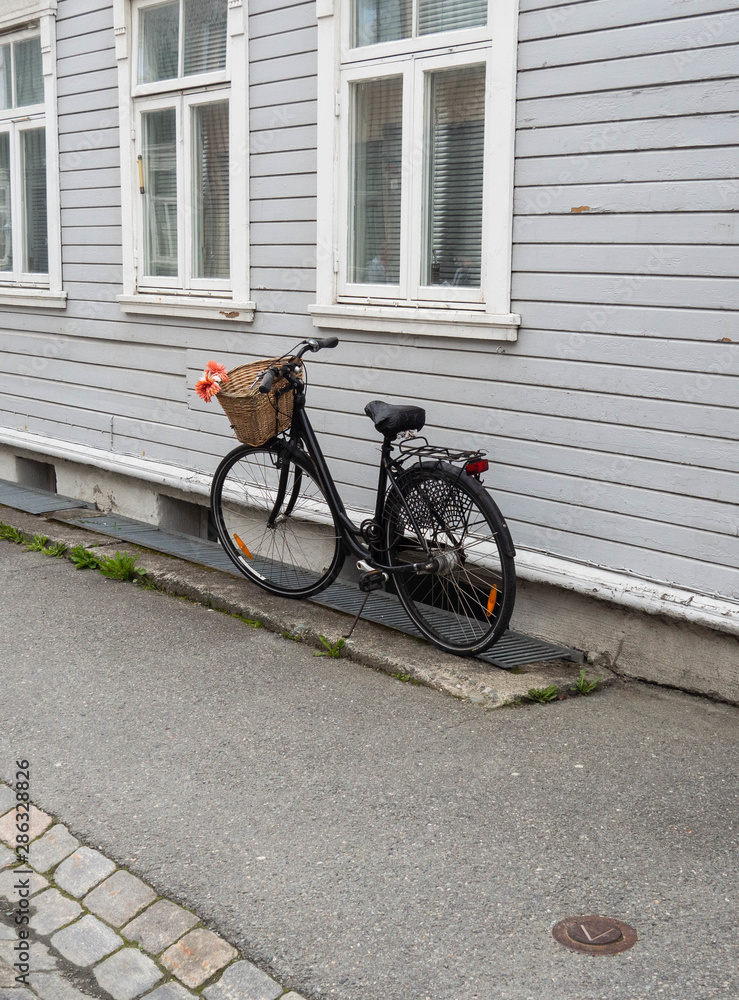 A black fashionable bicycle with a basket on the handlebars stands against the wall of an old wooden white house on a paved sidewalk