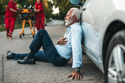 Elegant senior man with heart attack symptoms sitting on the road emergency medical service workers trying to help him. Driver assistance service.