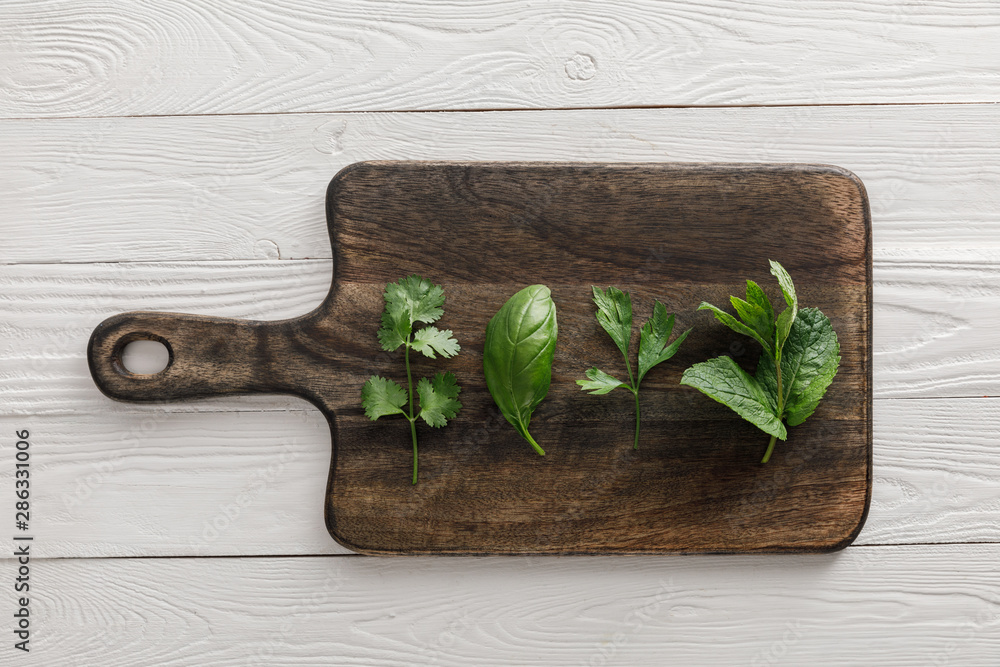 Top view of brown wooden cutting board with parsley, basil, cilantro and peppermint leaves on white surface