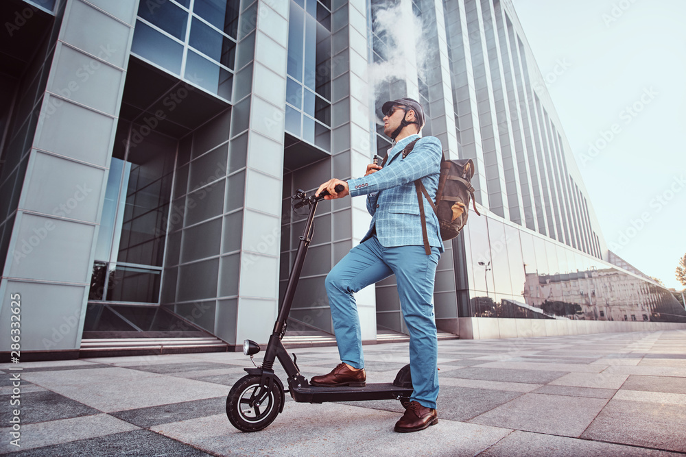 Pensive man in protective helmet and sunglasses is smoking vape while holding his electrical scooter. Pensive man in protective helmet and sunglasses is smoking vape while holding his electrical scooter.