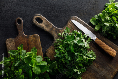 Top view of brown wooden cutting boards with parsley, peppermint and cilantro bundles with knife on dark surface